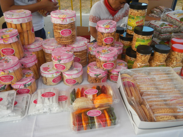 Eine Gruppe von Menschen um einen Tisch mit verschiedenen Lebensmitteln auf einem Food-Festival, mit einem Geländer und Straße im Hintergrund.
