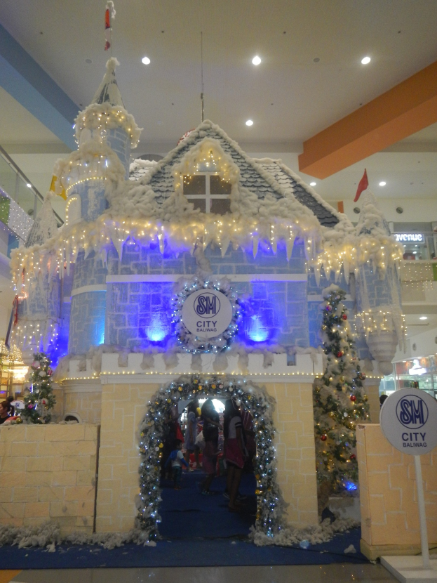 Ein festlich geschmücktes Weihnachtsdorf mit einer burgenähnlichen Struktur, umgeben von Menschen in einem Einkaufszentrum mit beleuchteter Decke und einer Tafel auf der rechten Seite.