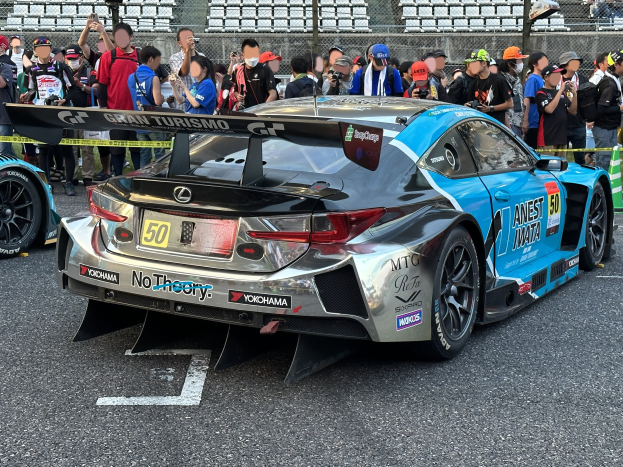 Ein schwarzer Lexus RC F GT3 Rennauto umgeben von Zuschauern mit Kameras und Handys, hinter Maschendrahtzaun und verstreuten Stühlen.