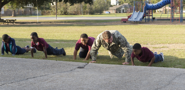 Kinder machen Liegestütze auf Gras in einem Park mit Spielplatzgeräten, Häusern, einem Auto, Bäumen und einem bewölkten Himmel.