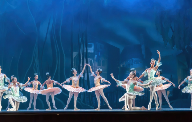 Eine Gruppe von Balletttänzerinnen in bunten Kostümen, die *Der Nussknacker* auf der Bühne des Royal Opera House tanzen, bewegen sich synchronisiert vor einem blauen Hintergrund.