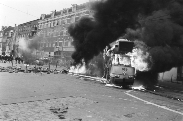 Schwarzes und weißes Foto eines brennenden Zuges auf einer Straße, mit Rauch, der aus ihm aufsteigt, Menschen auf dem Bürgersteig links und Gebäuden, Strommasten und Drähten im Hintergrund.