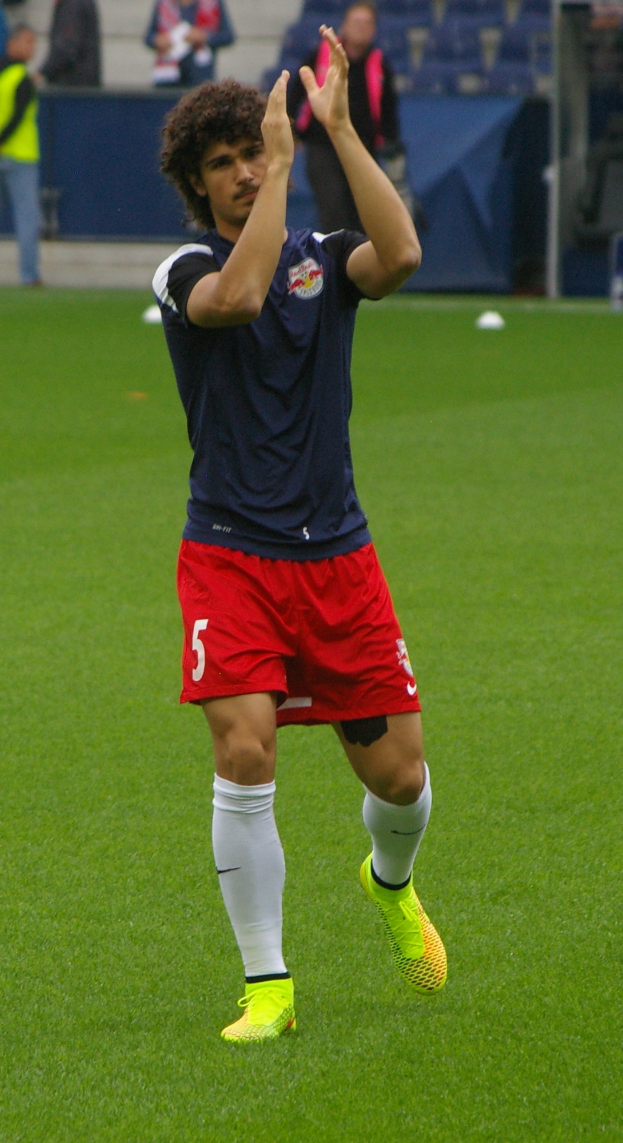 Ein Mann in einem blauen Hemd und roten Shorts steht auf einem Fußballfeld, umgeben von einer Gruppe von Menschen, bereit für ein Fußballspiel.