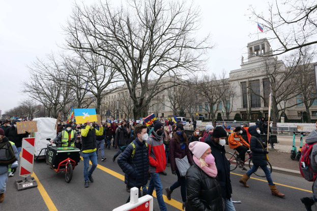 Schwarz-weißes Bild einer Protestdemonstration auf einem Schulgelände, mit Menschen, die Fahnen in einer Reihe auf einem Schotterweg halten, Bäume, Gebäude und einen klaren Himmel im Hintergrund.