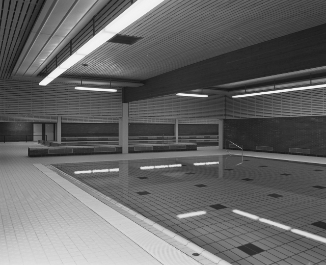 Indoor-Schwimmbad mit klarem Wasser, gefliestem Boden, Wänden und Deckenbeleuchtung in einem großen, ruhigen Gebäude.