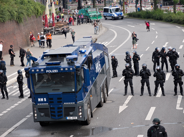Gruppe von Polizisten in Uniform vor einem gepanzerten Fahrzeug auf einer Stra├če stehend, umgeben von bewaffneten Personen mit Helmen, mit Geb├Ąuden, B├Ąumen und Verkehrszeichen im Hintergrund.