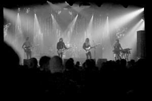 Ein Schwarz-Weiß-Bild eines Musikkonzerts mit vier Künstlern auf der Bühne: zwei Gitarristen und eine singende Frau, mit Zuschauern unten und Lichtern oben.