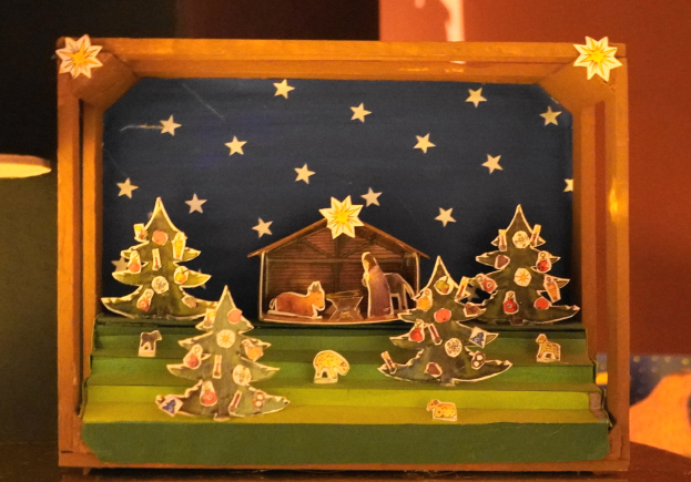 Eine Krippe in einer mit Weihnachtsbäumen, Tieren und einem Haus geschmückten Holzschachtel, die auf einem Tisch mit einer Lampe links daneben und einer Wand im Hintergrund steht.