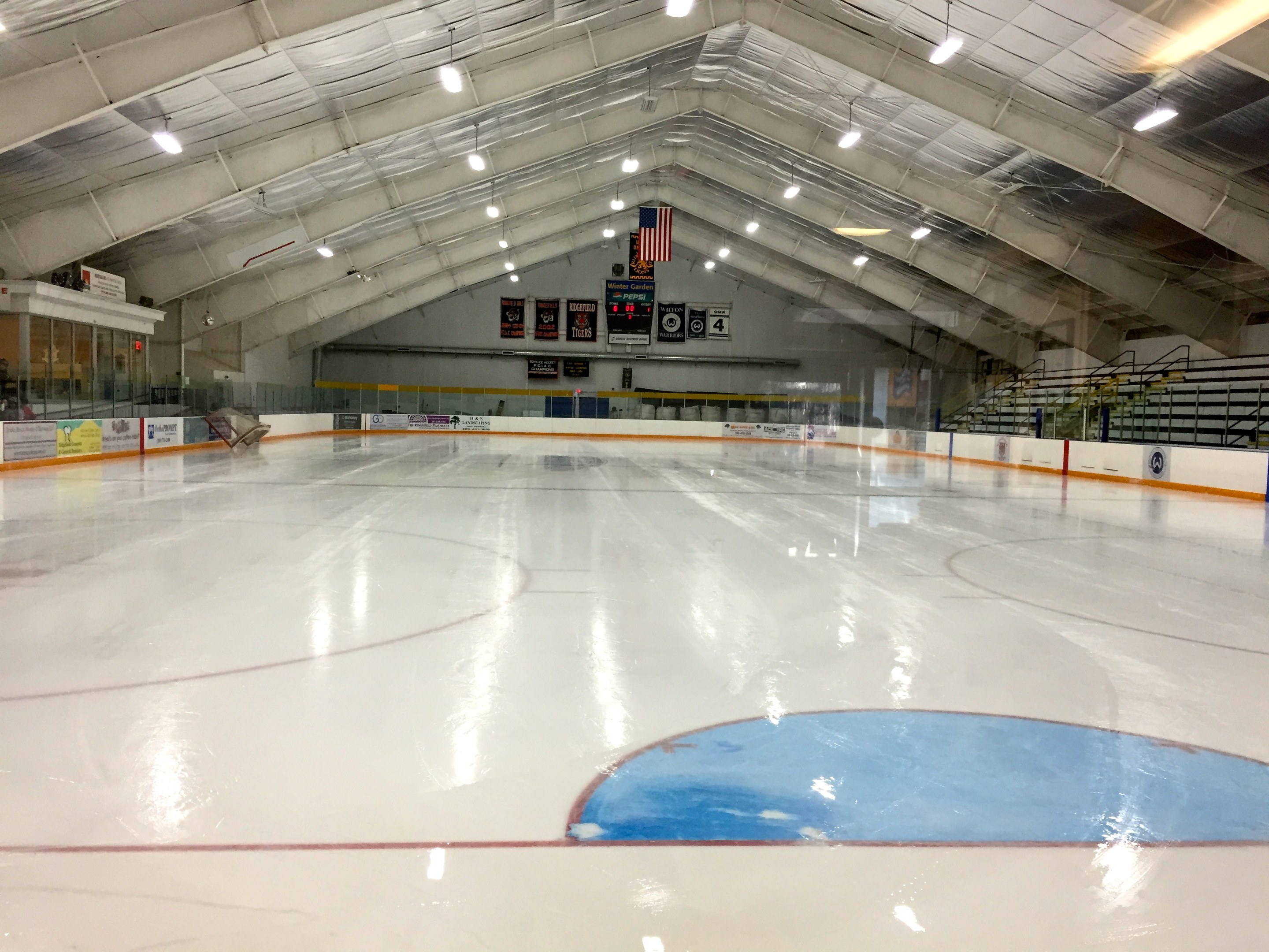 Großes Indoor-Eisstadion mit Deckenbeleuchtung, Werbetafeln, einer Wand, Treppen mit Geländern und einigen Menschen, was auf eine Sportanlage hinweist, die likely für Hockey genutzt wird.