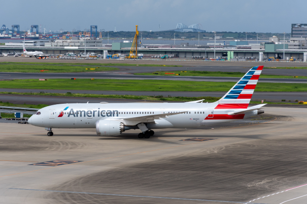 American Airlines-Flugzeug auf der Rollbahn eines Flughafens, umgeben von Gras, Fahrzeugen, Gebäuden, Masten, Kränen, Bäumen und einem klaren blauen Himmel.