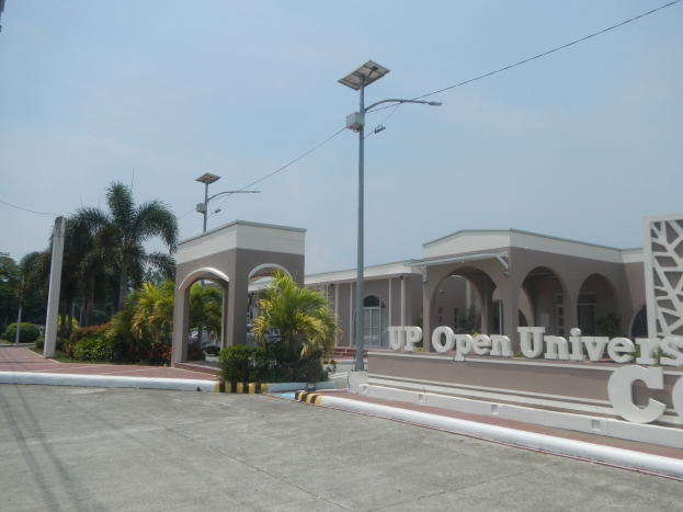 Offene Universitätscampus-Eingang mit einer Straße, die zu einem Gebäude führt, umgeben von Bäumen und Pflanzen, eine texttragende Wand auf der rechten Seite und Himmel im Hintergrund.