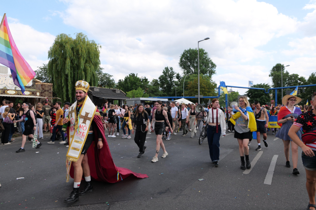 Eine Gruppe von Menschen marschiert bei der Gay Pride Parade 2018, einige tragen Musikinstrumente und andere Hüte, während sie eine Regenbogenflagge schwenken und eine Straße entlanggehen, die von Laternenpfählen, Bäumen, Schuppen und einem bewölkten Himmel gesäumt ist.