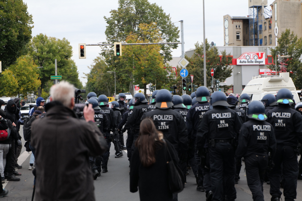 Eine Gruppe von Menschen geht eine Straße entlang, umgeben von Polizeibeamten, einige tragen Helme und tragen Taschen oder Kameras, mit Fahrzeugen, Verkehrszeichen, Schildern, Bäumen, Gebäuden und einem klaren Himmel im Hintergrund.