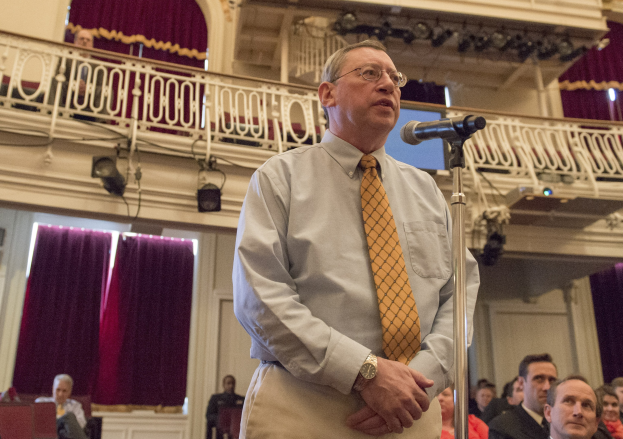 Ein Mann in Hemd, Krawatte und Brille spricht in ein Mikrofon an einem Rednerpult, vor einer sitzenden Menge mit Bühnenbeleuchtung und Vorhängen im Hintergrund.