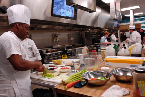 Eine Gruppe von Köchen in einer Küche bei der Zubereitung von Essen auf einem Schneidbrett, wobei ein Koch im Vordergrund in einer weißen Schürze und einer weißen Haube steht und ein Messer hält und von Utensilien, Schüsseln, Gemüse, einem Handy, Flaschen und anderen Gegenständen umgeben ist, während im Hintergrund ein Fernseher und Deckenlampen zu sehen sind.