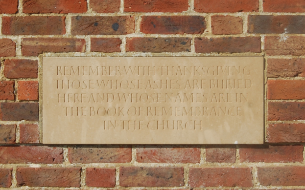 Eine Backsteinmauer mit einer Plakette, auf der steht: "Erinnere mit Dankbarkeit an diejenigen, deren Asche hier begraben ist und deren Namen im Buch der Erinnerung in der Kirche stehen."