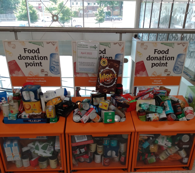 Orange Schübe mit Lebensmittelspenden, darunter Flaschen, Dosen und Kisten, neben Informationsschildern, mit Fahrzeugen, Bäumen, Gebäuden und Himmel durch Glasfenster sichtbar.