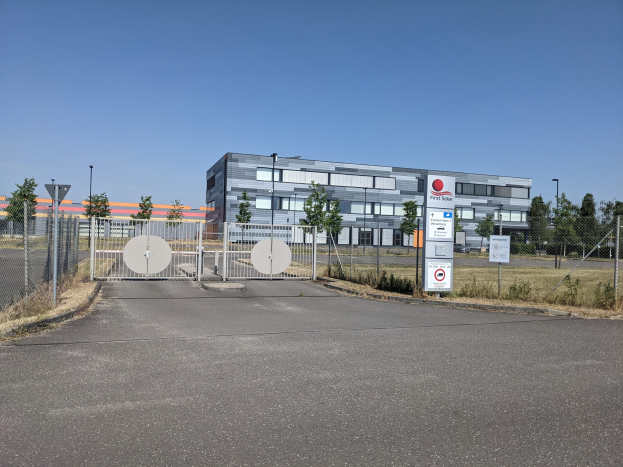 Großes industrielles Gebäude mit der Bezeichnung "First Solar" und zahlreichen Fenstern, umgeben von einem Metallzaun, umgeben von Bäumen, Pflanzen, Gras, Schildern, Straßenlaternen und einem Weg unter einem bewölkten Himmel.