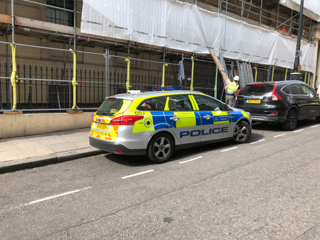 Ein Polizeiwagen steht an der Straßenseite neben einem Gebäude, zwei Helme tragende Individuen stehen in der Nähe; ein Geländer und ein Pfahl sind im Vordergrund, und ein weißer Stoff ist über das Gebäude drapiert, was auf eine polizeiliche Untersuchung nach einem Messerangriff in London hindeutet.