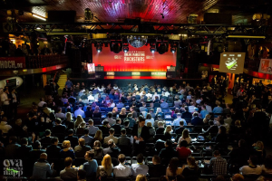 Eine große Menschenmenge sitzt auf Stühlen vor einer Bühne mit Bildschirmen und Bannern, beleuchtet von Scheinwerfern und umgeben von Absperrungen, voller Erwartung auf den Beginn eines Online-Marketing-Rockstars-Events, mit einem Wasserzeichen in der linken unteren Ecke.