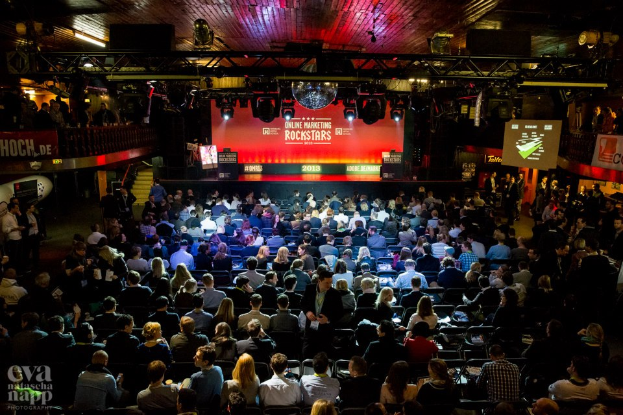 Eine große Menschenmenge sitzt auf Stühlen vor einer Bühne mit Bildschirmen und Bannern, beleuchtet von Scheinwerfern und umgeben von Absperrungen, voller Erwartung auf den Beginn eines Online-Marketing-Rockstars-Events, mit einem Wasserzeichen in der linken unteren Ecke.