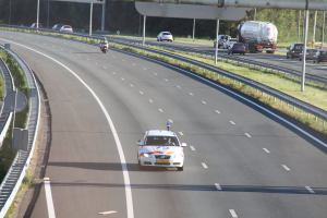 Ein Auto fährt auf einer Autobahn mit einem Polizeiwagen am Straßenrand, anderen Fahrzeugen, Geländern, Pfählen, Brettern und Bäumen im Hintergrund.