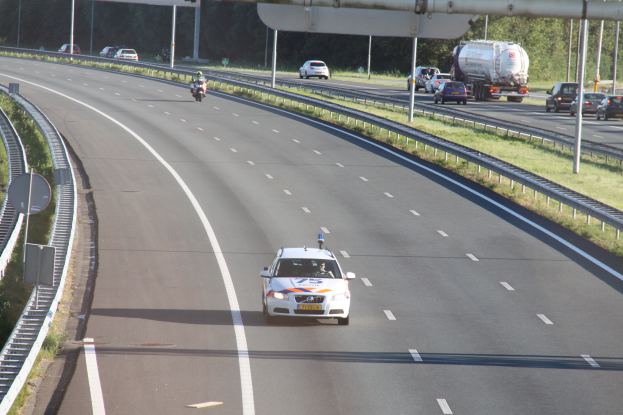 Ein Auto fährt auf einer Autobahn mit einem Polizeiwagen am Straßenrand, anderen Fahrzeugen, Geländern, Pfählen, Brettern und Bäumen im Hintergrund.