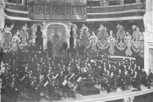 Ein altes Schwarz-Weiß-Foto eines großen Orchesters in einem Auditorium, mit Musikern auf der Bühne sitzend und stehend, einem Podium in der Mitte und Skulpturen an den Wänden im Hintergrund.