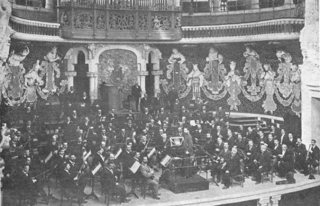 Ein altes Schwarz-Weiß-Foto eines großen Orchesters in einem Auditorium, mit Musikern auf der Bühne sitzend und stehend, einem Podium in der Mitte und Skulpturen an den Wänden im Hintergrund.