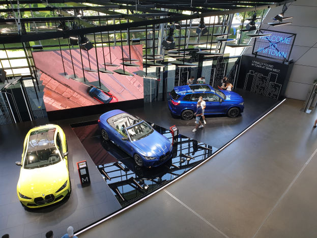Eine Gruppe von Menschen steht im BMW Museum in München, mit Autos zur Linken und einem Schild mit der Aufschrift "BMW Museum München" zur Rechten, mit Blick auf Bäume und Gebäude durch Glasfenster.