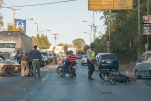 Eine Gruppe von Menschen steht um ein verunglücktes Motorrad am Straßenrand mit mehreren Fahrzeugen, darunter ein Lastwagen, und einem Hintergrund aus Bäumen, Polen, Lampen und Schildern unter dem Himmel.