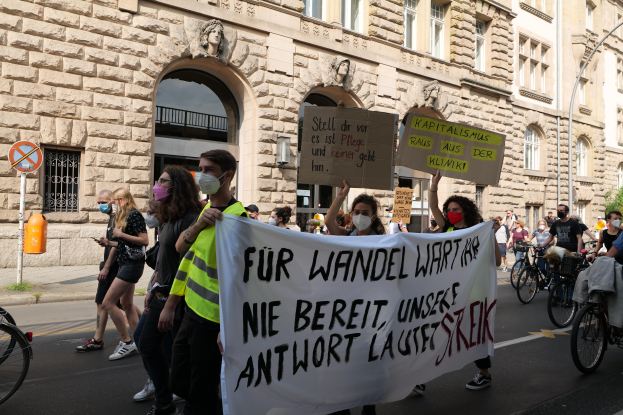 Eine Gruppe von Menschen marschiert auf der Straße in Berlin, hält Schilder und Banner hoch und einige fahren Fahrräder, vor einem Gebäude mit Bögen, Säulen, Skulpturen und Bäumen.