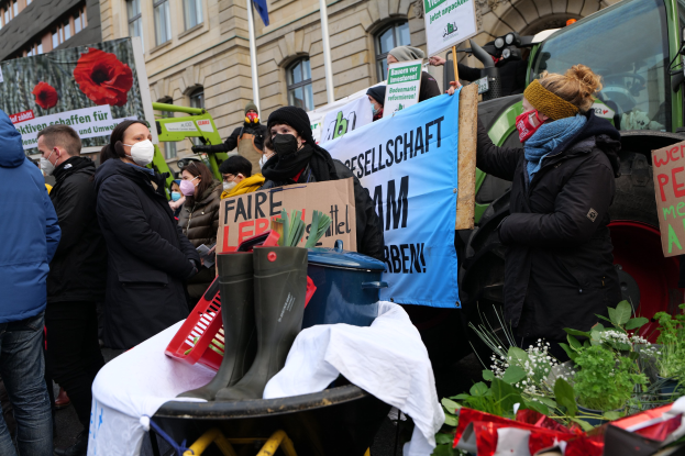 Maskierte Demonstranten mit Protestschildern vor einem Lastwagen mit einem Tisch, Pflanzen und einem Gebäude mit Flagge im Hintergrund.