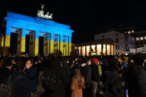 Eine Menschenmenge steht vor dem Brandenburger Tor in Berlin, Deutschland, viele tragen Mützen und Taschen, einige halten Schilder, mit den Säulen und Statuen des Tors im Hintergrund und Gebäuden mit Fenstern im Hintergrund unter einem dunklen Himmel.