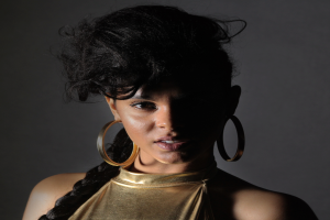 Eine Frau in einem goldenen Kleid und großen Creolen steht selbstbewusst vor einer Wand, ihr glatter Bob-Haarschnitt und natürliches Make-up unterstreichen ihren entschlossenen Gesichtsausdruck, während sie direkt in die Kamera schaut.