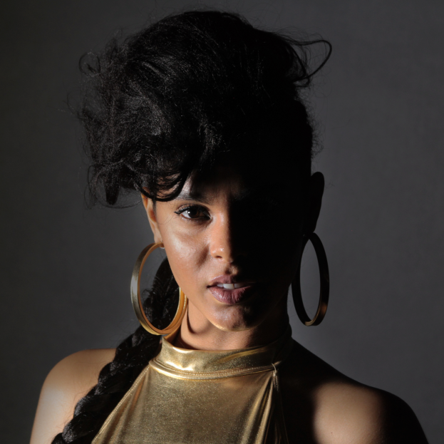 Eine Frau in einem goldenen Kleid und großen Creolen steht selbstbewusst vor einer Wand, ihr glatter Bob-Haarschnitt und natürliches Make-up unterstreichen ihren entschlossenen Gesichtsausdruck, während sie direkt in die Kamera schaut.