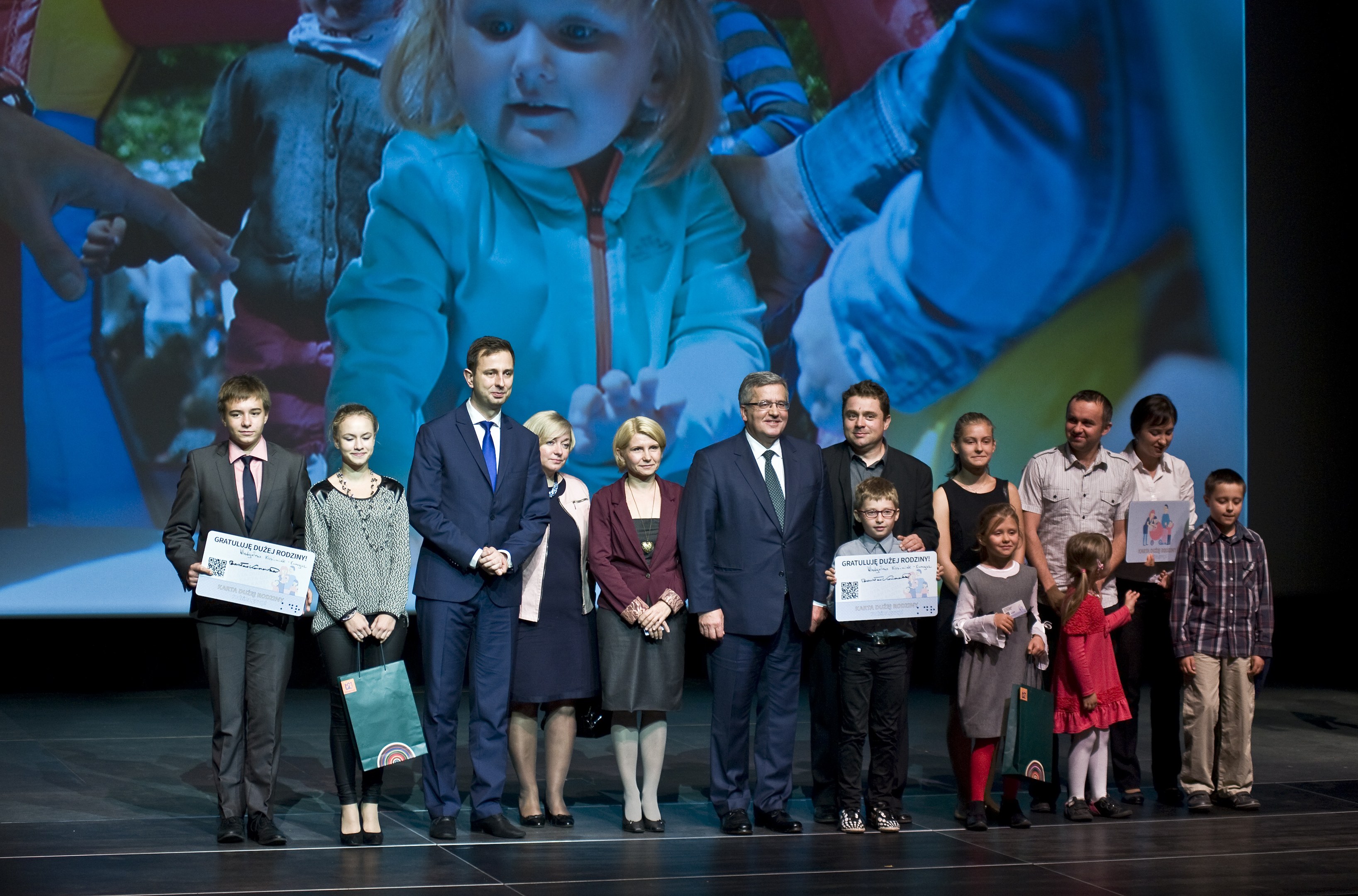 Gruppe von Menschen auf einer Bühne mit Taschen und Urkunden, im Hintergrund ein Bildschirm mit einem Kind und anderen Personen.