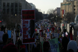 Eine Gruppe von Menschen protestiert auf einer Straße, hält Plakate in den Händen, mit Gebäuden, Bäumen, Laternenmasten und dem Himmel im Hintergrund.