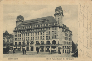 Eine alte Postkarte, die das Hotel Handelshof in Rühmann, Deutschland, mit einem großen Gebäude, Menschen, Fahrzeugen, Bäumen und einem bewölkten Himmel sowie Text zeigt.