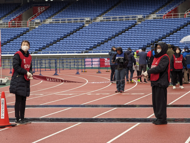 Gruppe von Menschen auf einer Rennbahn, die ein Band an der Startlinie halten, mit einem Verkehrskegel links und Event-Aufbau im Hintergrund.