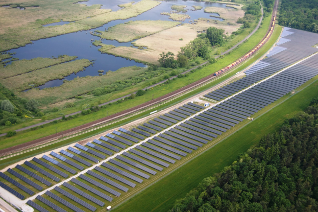 Luftaufnahme eines Solarparks mit Panelen in einem Feld, umgeben von Bäumen, Gras, Wasser und einer nahen Bahnlinie.