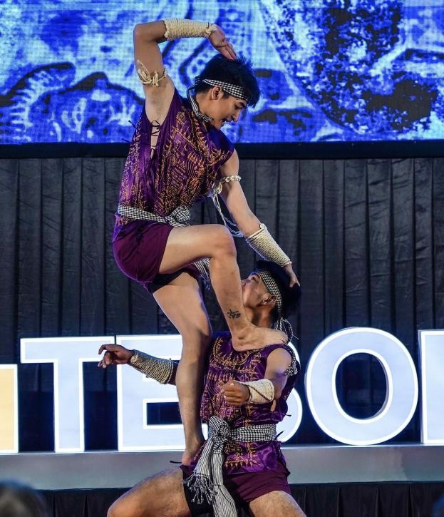 Zwei Akrobaten performen auf der Bühne, der Mann hebt die Frau in lila Kleidern hoch, während das Publikum unten zusieht.