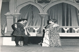 Ein Schwarz-Weiß-Foto eines Mannes und einer Frau, die auf einer Bühne vor einer Wand mit Säulen und einem Vorhang im Hintergrund stehen, beide in Abendgarderobe gekleidet.