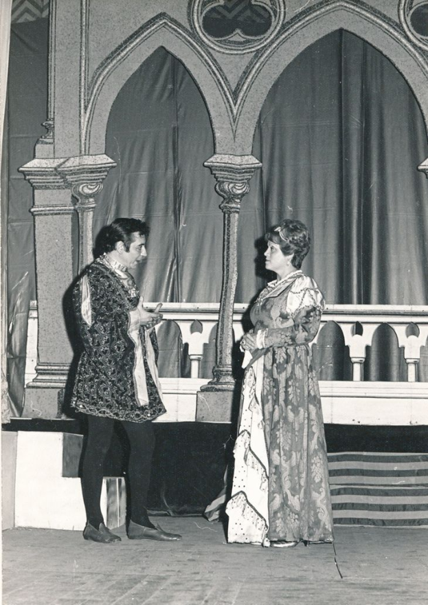 Ein Schwarz-Weiß-Foto eines Mannes und einer Frau, die auf einer Bühne vor einer Wand mit Säulen und einem Vorhang im Hintergrund stehen, beide in Abendgarderobe gekleidet.