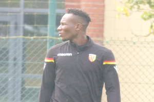 Ein Mann in einer schwarzen Jacke steht auf einem Fussballfeld, umgeben von einem Zaun und einem Baum auf der rechten Seite, mit einem entschlossenen Gesichtsausdruck.