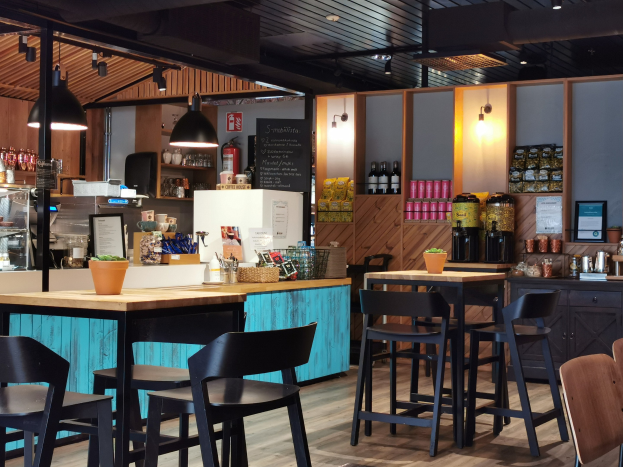 Innenraum eines Restaurants mit Holztischen und -stühlen, einem Topf mit einer Pflanze auf einem Tisch, einer Schiefertafel, einem Kühlschrank, einem Feuerlöscher, Regalen mit Flaschen, Schränken mit Gegenständen, einem gerahmten Foto an der Wand, Deckenlampen und Lampen.