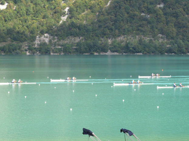 Eine Gruppe von Menschen, die bei einem Wettbewerb auf einem Gewässer rudern, umgeben von Bäumen und einem Hügel im Hintergrund, und Kleidung über Tribünen am Wasserrand drapiert.