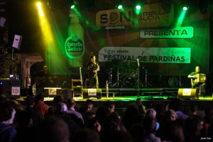 Eine Bühne mit Musikern, die Instrumente spielen, Lautsprechern, Mikrofonen, Werbetafeln und fokussiertem Licht, mit einer Flasche und anderen Gegenständen im Hintergrund.