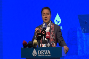 Ein Mann in Mantel und Schal steht an einem Podium mit Mikrofonen, ein Schild mit der Aufschrift "deva-Geschäftsführer sagt, es ist keine gute Idee für die Zukunft", ein Objekt und ein Bildschirm im Hintergrund.