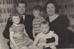 Eine Familie mit fünf Personen, darunter ein Mann in Anzug und Krawatte auf der linken Seite, eine Frau auf der rechten Seite und drei Kinder, die alle lächeln.